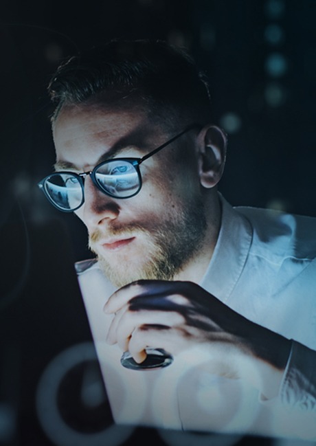 Person in glasses lit by a screen, hand on chin, concentrating on a laptop in a dark room