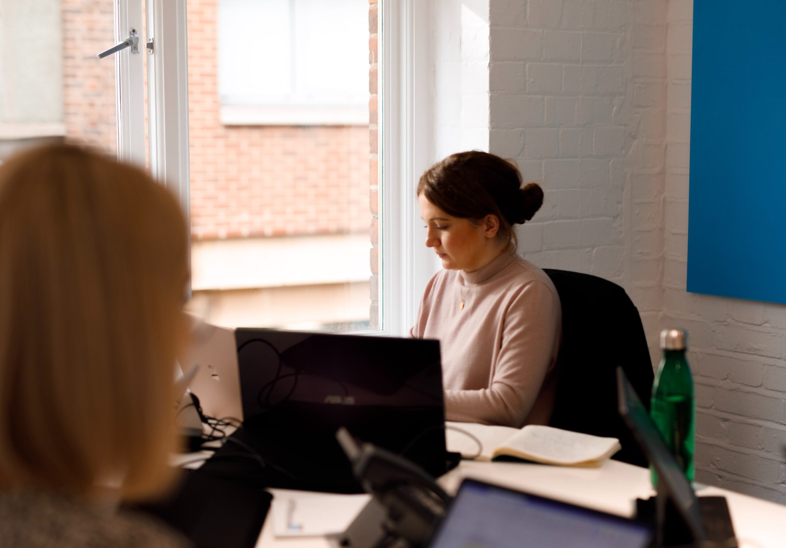 Colleagues working on laptops at a desk by a window in The Media People office