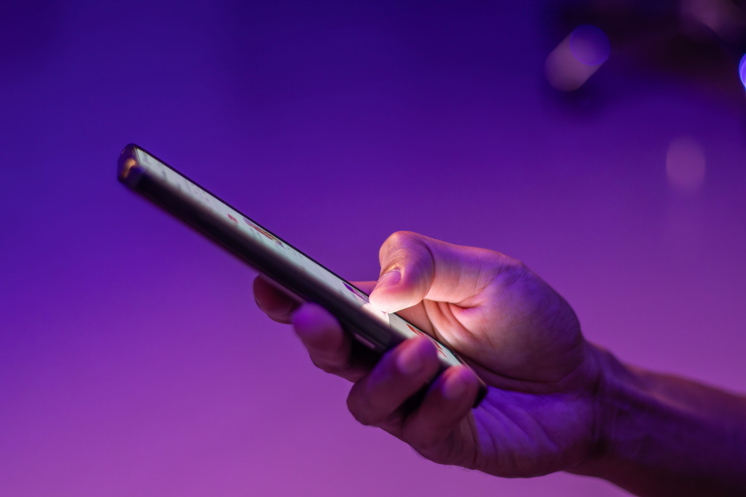Close-up of a hand scrolling on a smartphone in purple and blue neon light