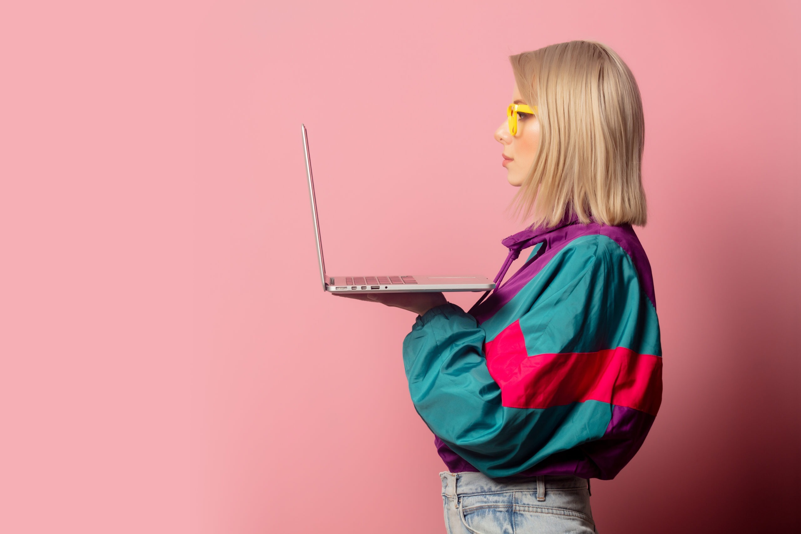 Person in a retro 90s windbreaker and yellow glasses holds an open laptop against a pink background