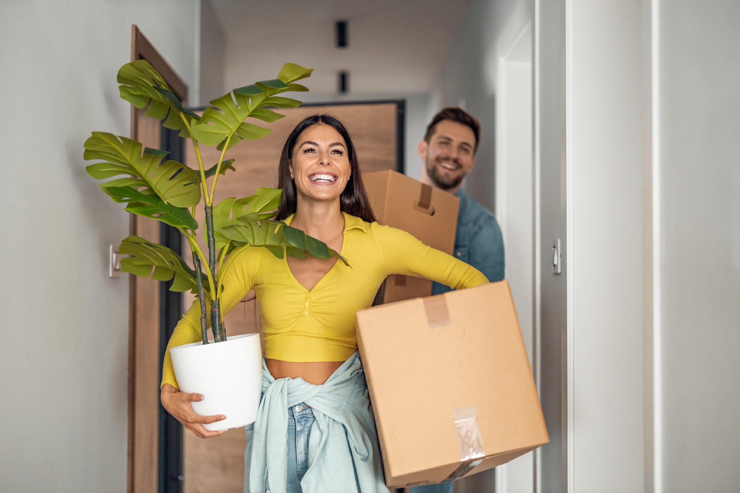 Two people smiling as they carry a potted plant and cardboard boxes into their new home