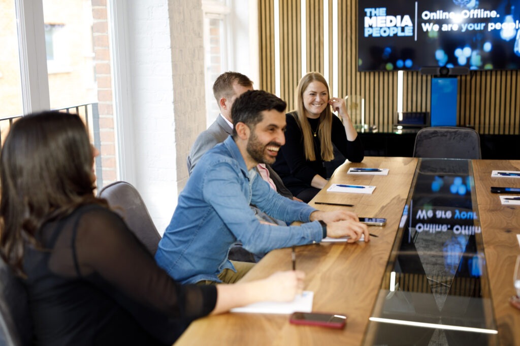 Colleagues laughing at a boardroom table, below a screen reading 'Online. Offline. We are your people.'