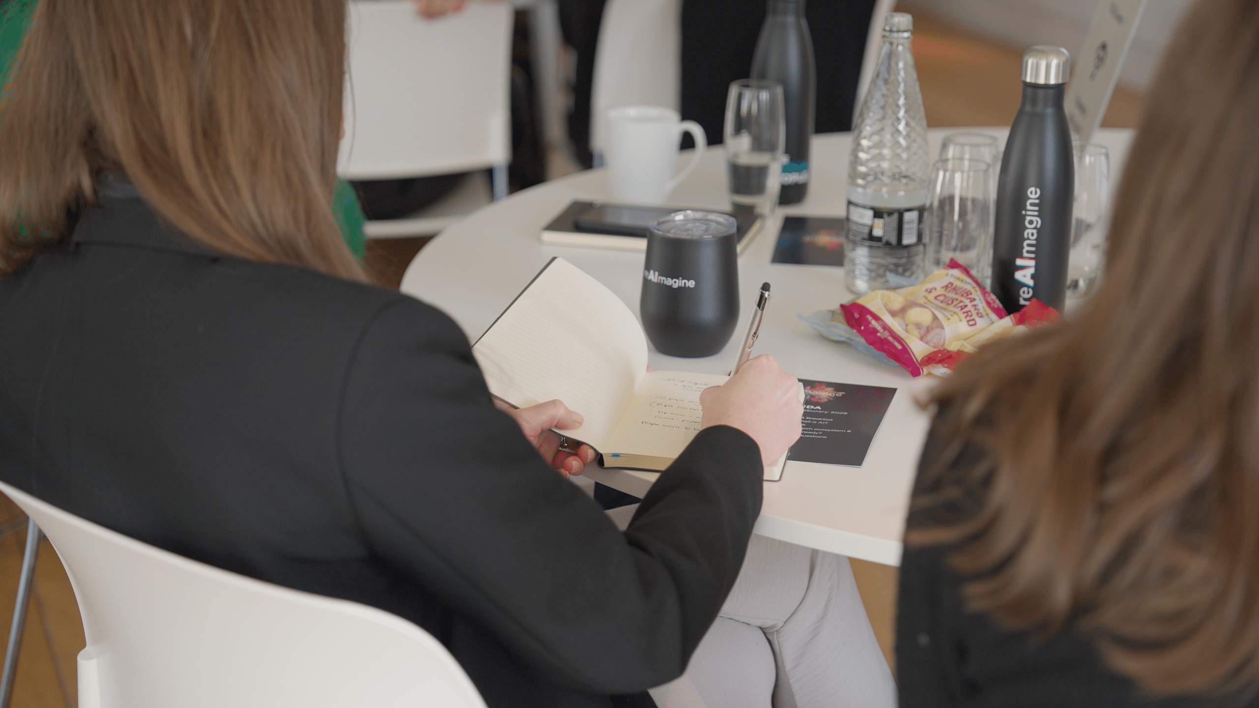 An attendee writes in a notebook at a table with reAImagine-branded bottles and cups at The Media People's AI Summit
