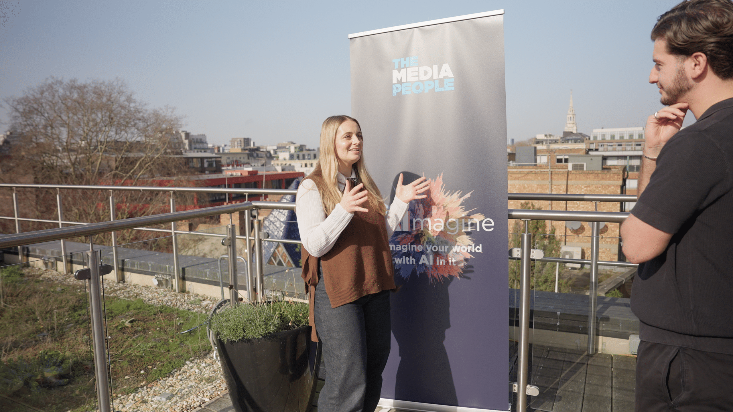 A person smiles while being interviewed on a sunny rooftop beside The Media People's reAImagine banner