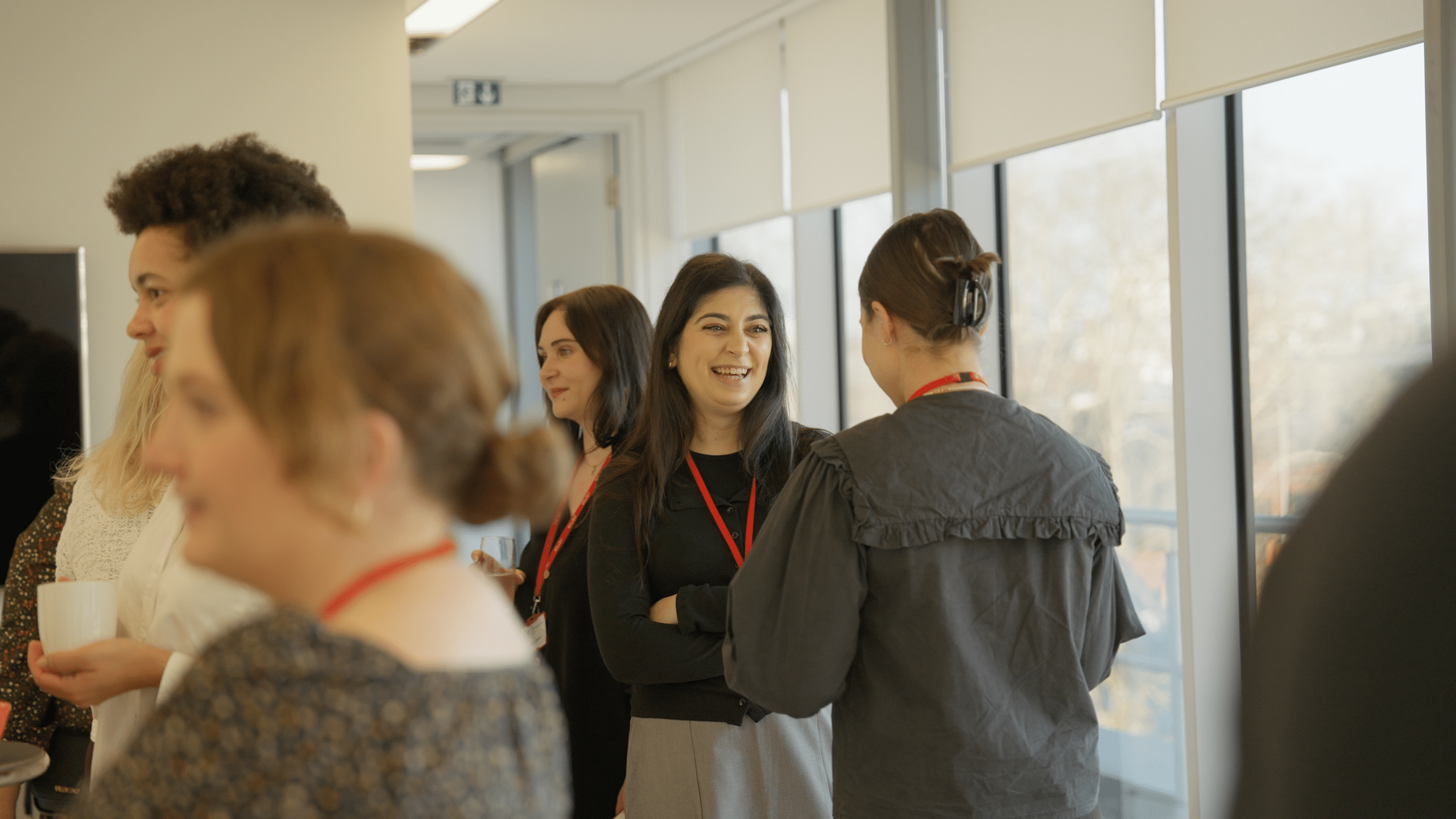 Guests wearing red lanyards chat and laugh beside tall windows at an office networking event