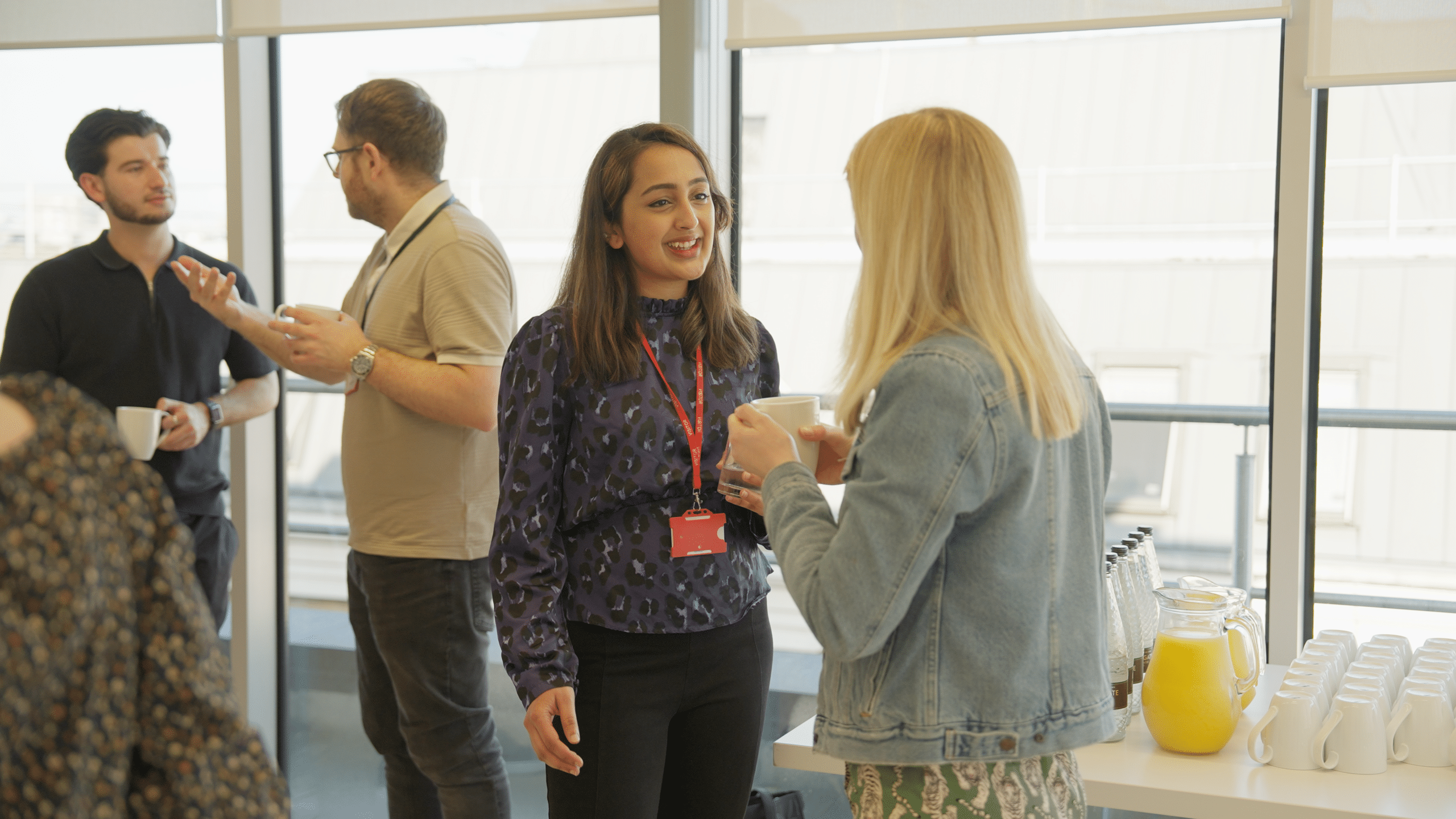 Guests in lanyards chat over coffee by a window, beside a table of orange juice and cups