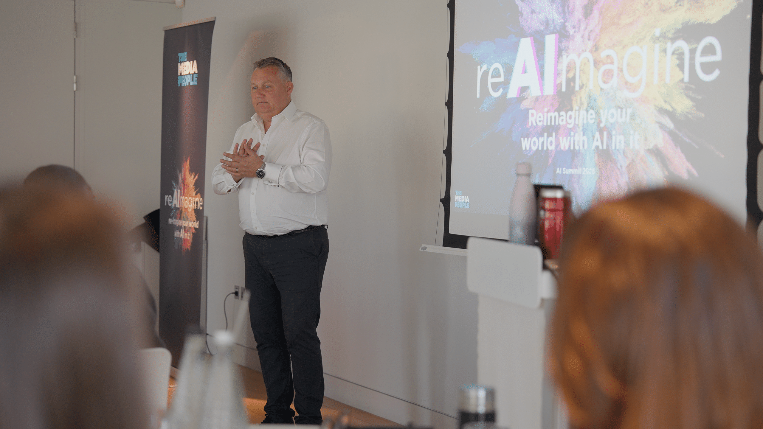 A speaker stands beside The Media People banner and a 'reAImagine: Reimagine your world with AI in it' screen