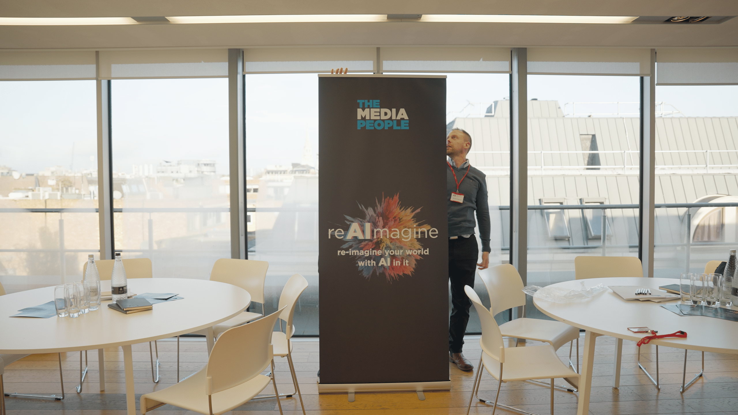 A person sets up The Media People's reAImagine banner in a bright event room with round tables and rooftop views
