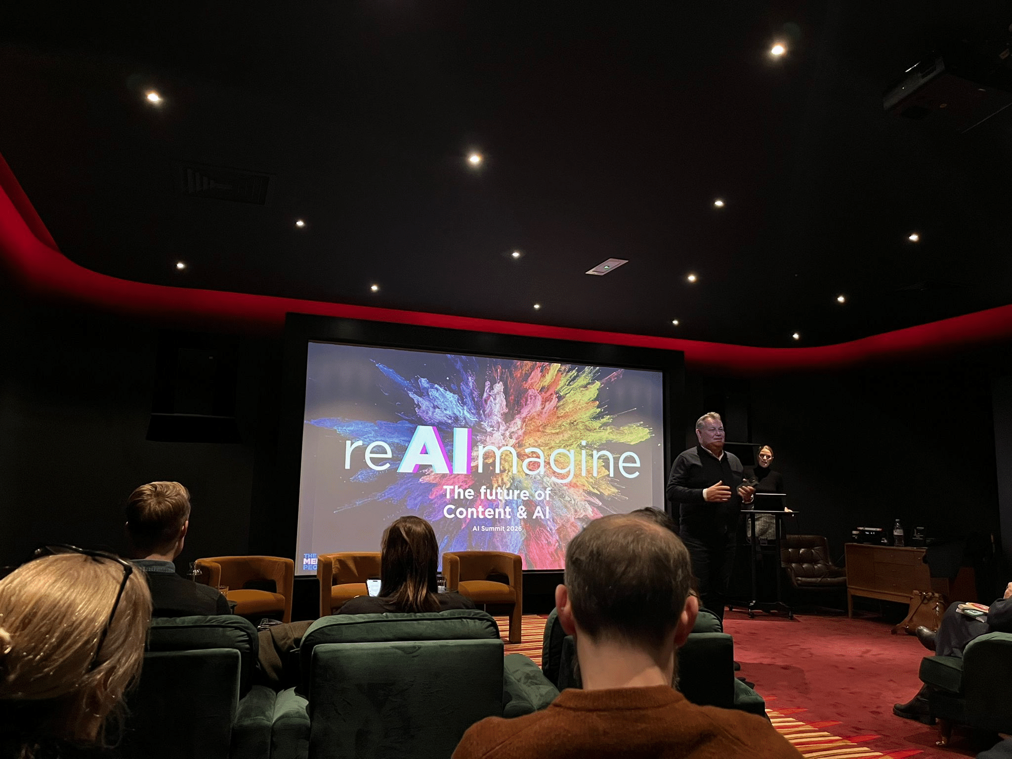 Speaker addresses an audience at the reAImagine AI Summit 2026, with the title slide on screen